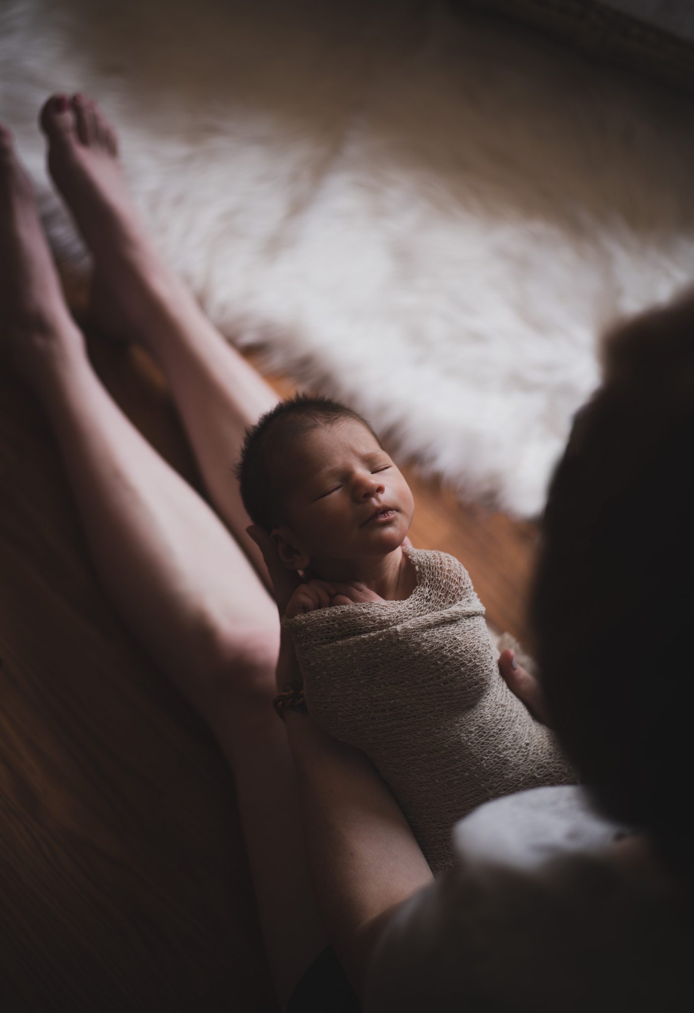 Dark and moody portrait of a newborn baby boy in the arms of his mother in their home in Hesperia California. Mother and baby both look happy as they pose in a lifestyle photoshoot for the inland empires number one family photographer. Baby boy is asian with fuzzy black hair in a child’s lifestyle photo shoot with a female photographer in high desert