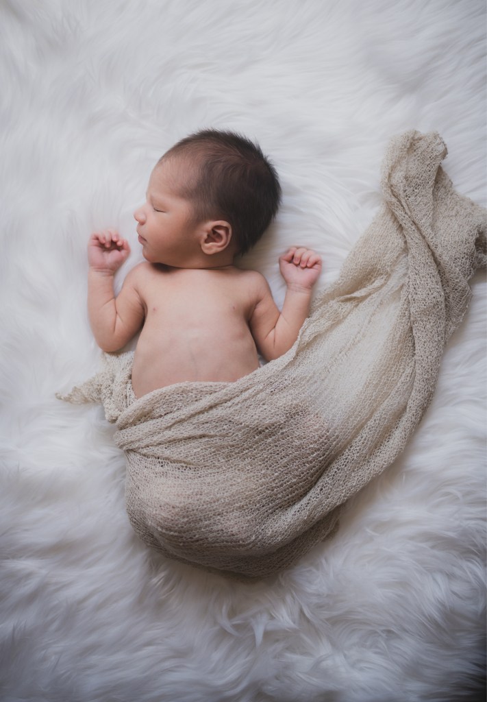 Dark and moody portrait of a newborn baby boy in the arms of his mother in their home in Hesperia California. Mother and baby both look happy as they pose in a lifestyle photoshoot for the inland empires number one family photographer. Baby boy is asian with fuzzy black hair in a child’s lifestyle photo shoot with a female photographer in high desert