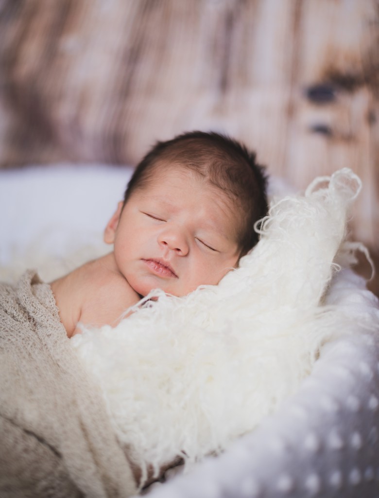 Dark and moody portrait of a newborn baby boy in the arms of his mother in their home in Hesperia California. Mother and baby both look happy as they pose in a lifestyle photoshoot for the inland empires number one family photographer. Baby boy is asian with fuzzy black hair in a child’s lifestyle photo shoot with a female photographer in high desert