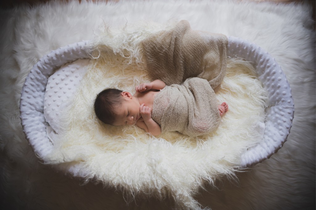 Dark and moody portrait of a newborn baby boy in the arms of his mother in their home in Hesperia California. Mother and baby both look happy as they pose in a lifestyle photoshoot for the inland empires number one family photographer. Baby boy is asian with fuzzy black hair in a child’s lifestyle photo shoot with a female photographer in high desert