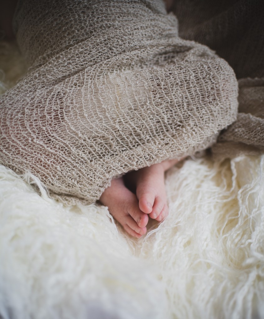Dark and moody portrait of a newborn baby boy in the arms of his mother in their home in Hesperia California. Mother and baby both look happy as they pose in a lifestyle photoshoot for the inland empires number one family photographer. Baby boy is asian with fuzzy black hair in a child’s lifestyle photo shoot with a female photographer in high desert