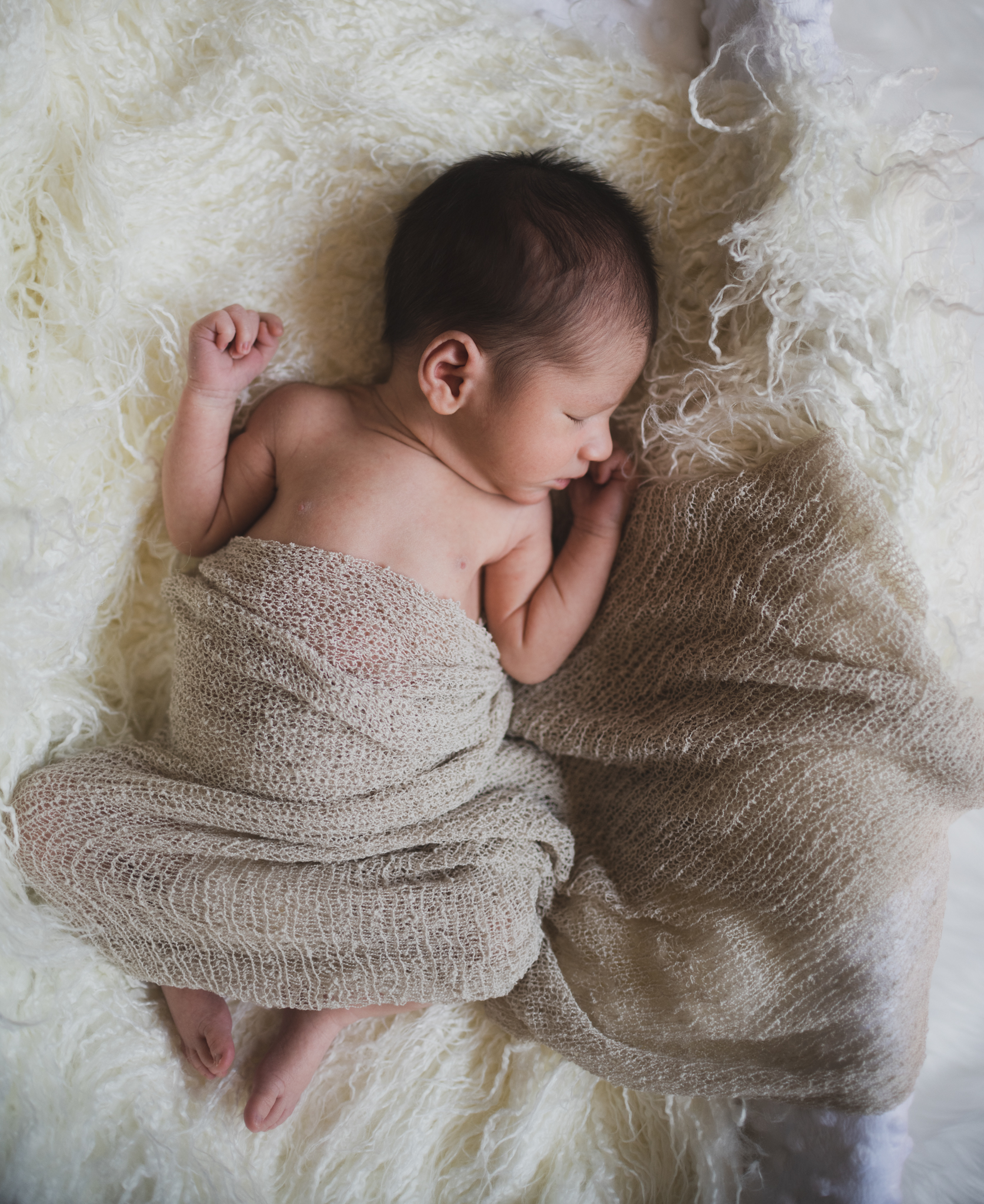 Dark and moody portrait of a newborn baby boy in the arms of his mother in their home in Hesperia California. Mother and baby both look happy as they pose in a lifestyle photoshoot for the inland empires number one family photographer. Baby boy is asian with fuzzy black hair in a child’s lifestyle photo shoot with a female photographer in high desert