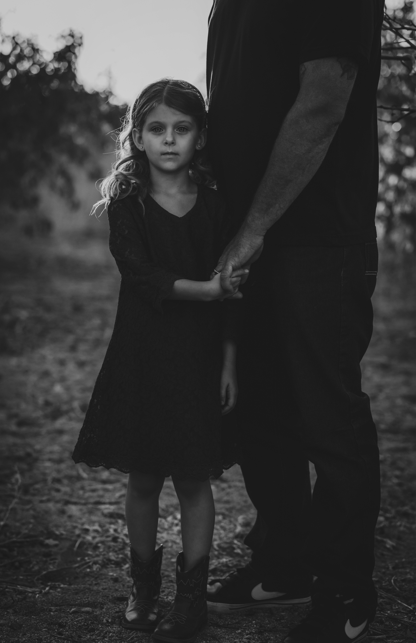 Dark and moody portrait of a family in a dead peach orchard posing for a family photoshoot in the high desert. There are rows of trees and a deep orange sunset setting behind the mountains of the inland empire. The family is wearing dark clothes smiling for the high deserts best family photographer
