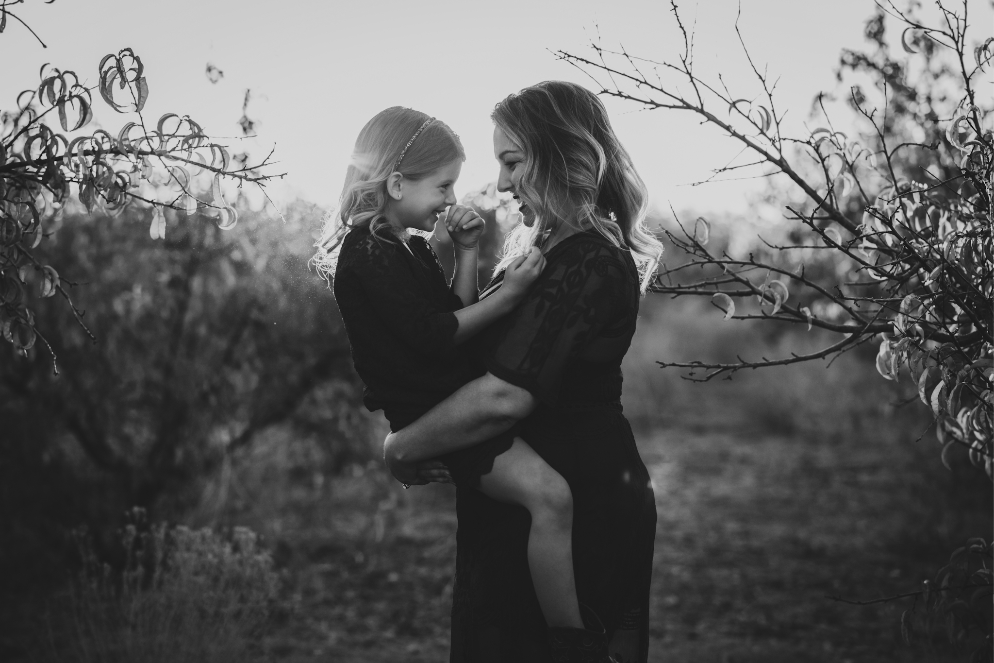 Dark and moody portrait of a family in a dead peach orchard posing for a family photoshoot in the high desert. There are rows of trees and a deep orange sunset setting behind the mountains of the inland empire. The family is wearing dark clothes smiling for the high deserts best family photographer