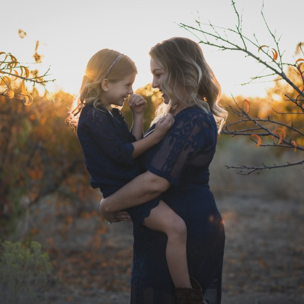 Dark and moody portrait of a family in a dead peach orchard posing for a family photoshoot in the high desert. There are rows of trees and a deep orange sunset setting behind the mountains of the inland empire. The family is wearing dark clothes smiling for the high deserts best family photographer