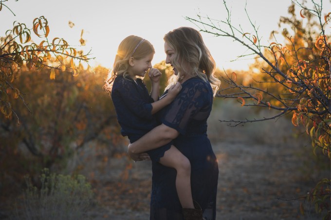 Dark and moody portrait of a family in a dead peach orchard posing for a family photoshoot in the high desert. There are rows of trees and a deep orange sunset setting behind the mountains of the inland empire. The family is wearing dark clothes smiling for the high deserts best family photographer
