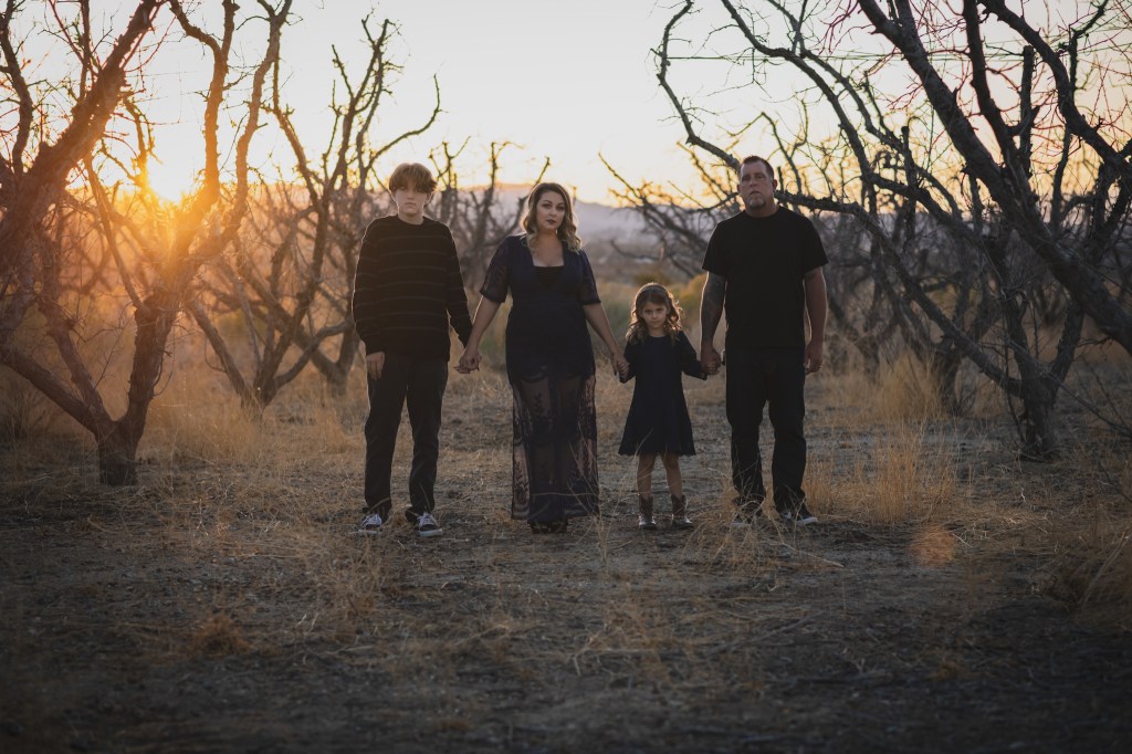 Dark and moody portrait of a family in a dead peach orchard posing for a family photoshoot in the high desert. There are rows of trees and a deep orange sunset setting behind the mountains of the inland empire. The family is wearing dark clothes smiling for the high deserts best family photographer