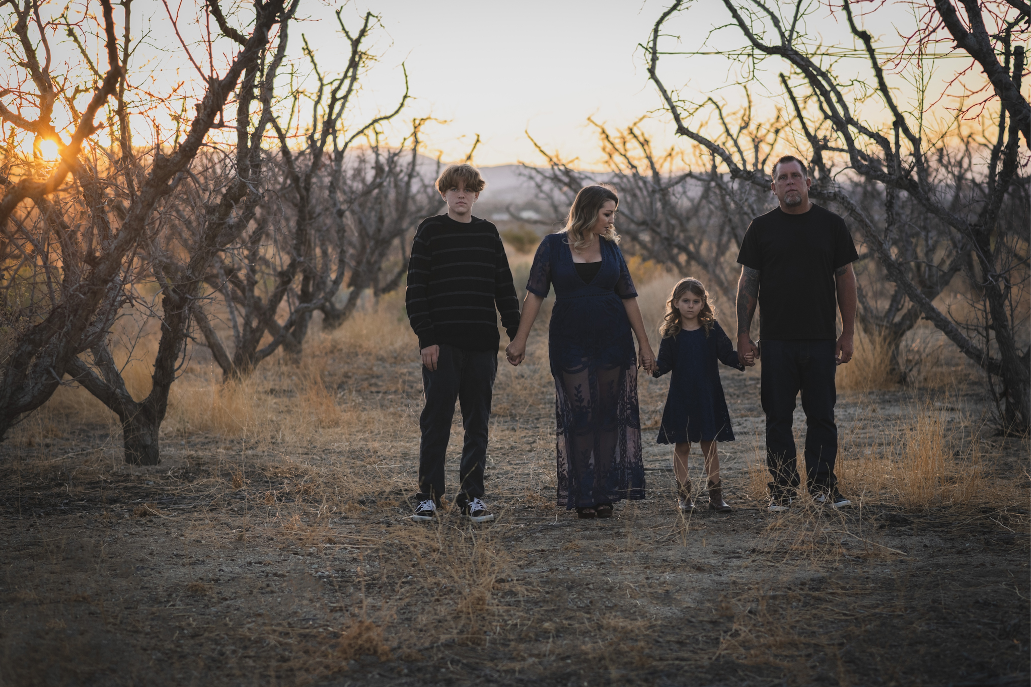 Dark and moody portrait of a family in a dead peach orchard posing for a family photoshoot in the high desert. There are rows of trees and a deep orange sunset setting behind the mountains of the inland empire. The family is wearing dark clothes smiling for the high deserts best family photographer