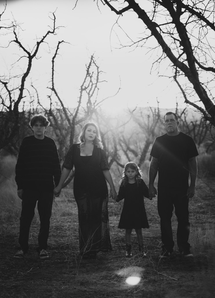 Dark and moody portrait of a family in a dead peach orchard posing for a family photoshoot in the high desert. There are rows of trees and a deep orange sunset setting behind the mountains of the inland empire. The family is wearing dark clothes smiling for the high deserts best family photographer