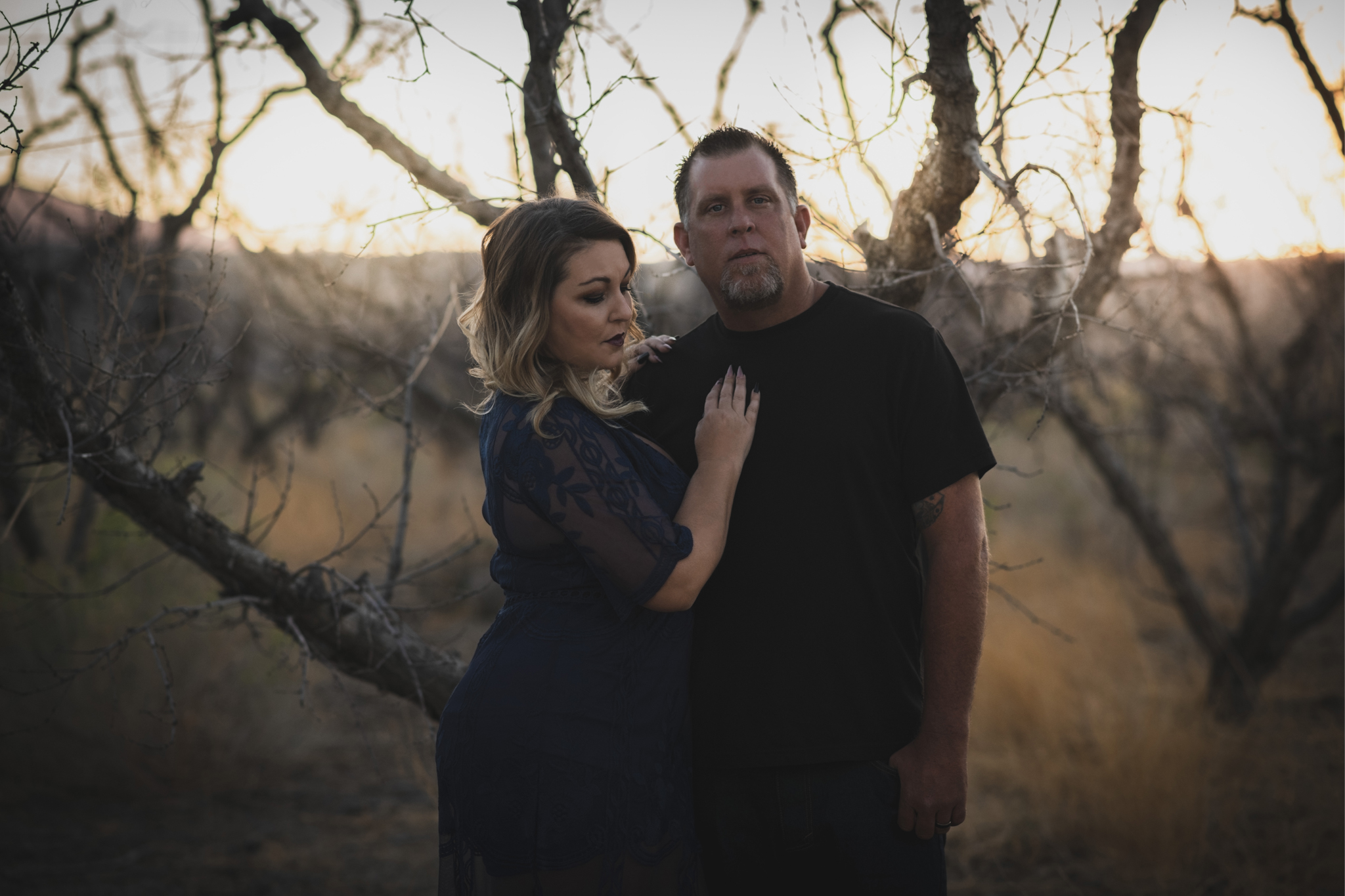 Dark and moody portrait of a family in a dead peach orchard posing for a family photoshoot in the high desert. There are rows of trees and a deep orange sunset setting behind the mountains of the inland empire. The family is wearing dark clothes smiling for the high deserts best family photographer