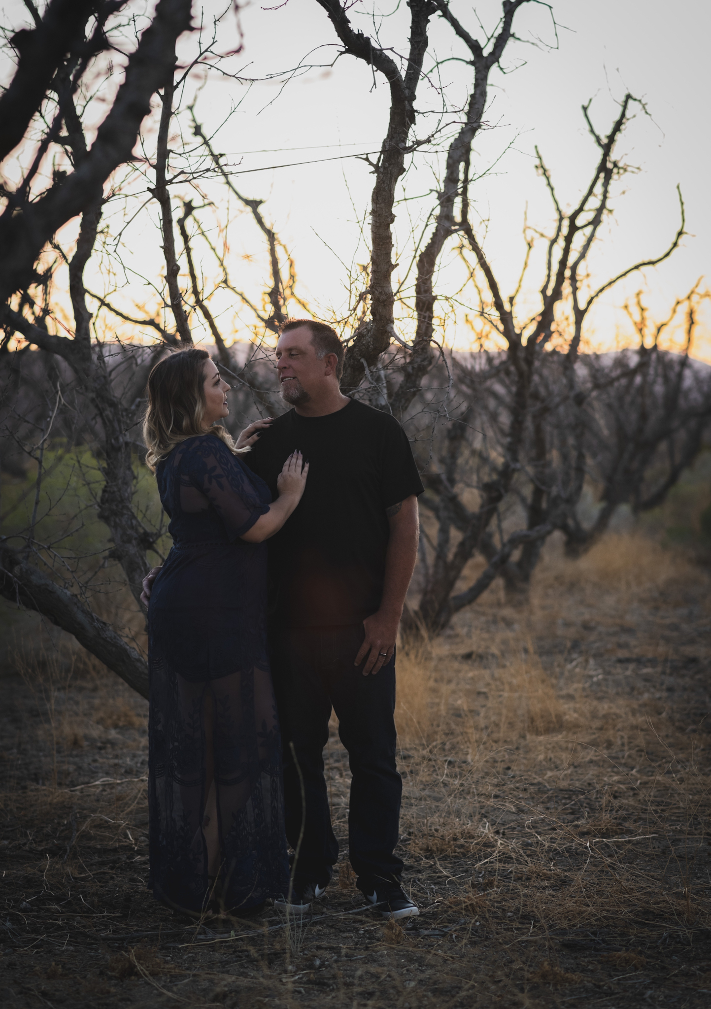 Dark and moody portrait of a family in a dead peach orchard posing for a family photoshoot in the high desert. There are rows of trees and a deep orange sunset setting behind the mountains of the inland empire. The family is wearing dark clothes smiling for the high deserts best family photographer