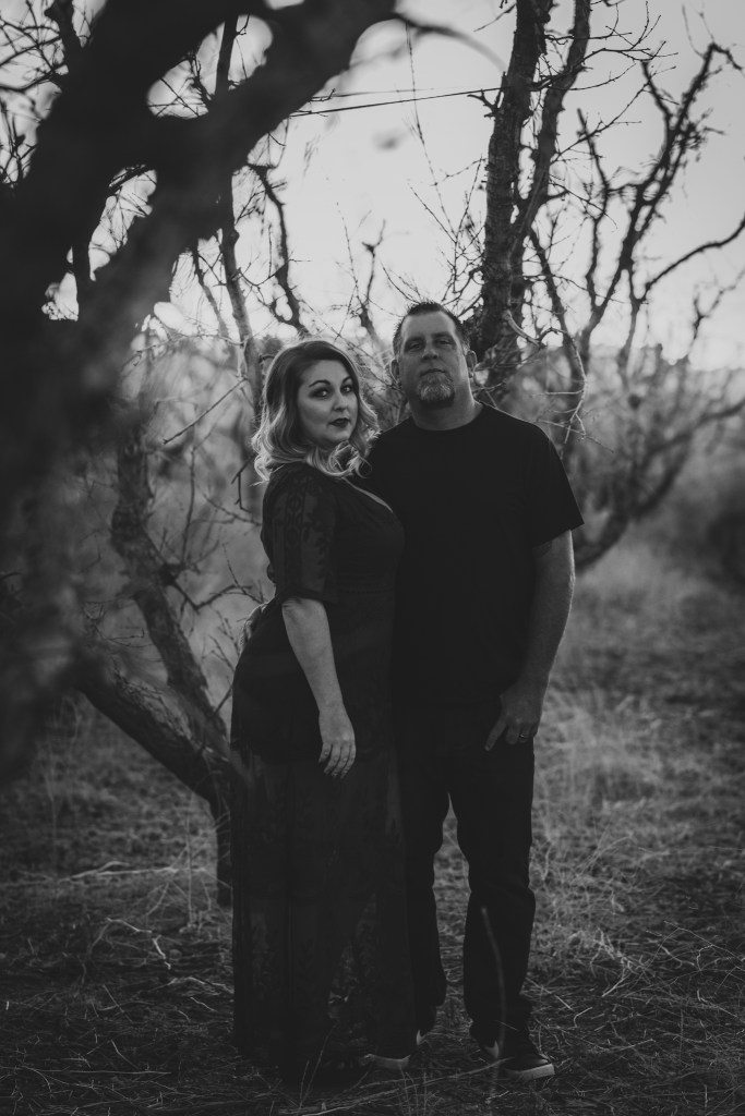 Dark and moody portrait of a family in a dead peach orchard posing for a family photoshoot in the high desert. There are rows of trees and a deep orange sunset setting behind the mountains of the inland empire. The family is wearing dark clothes smiling for the high deserts best family photographer