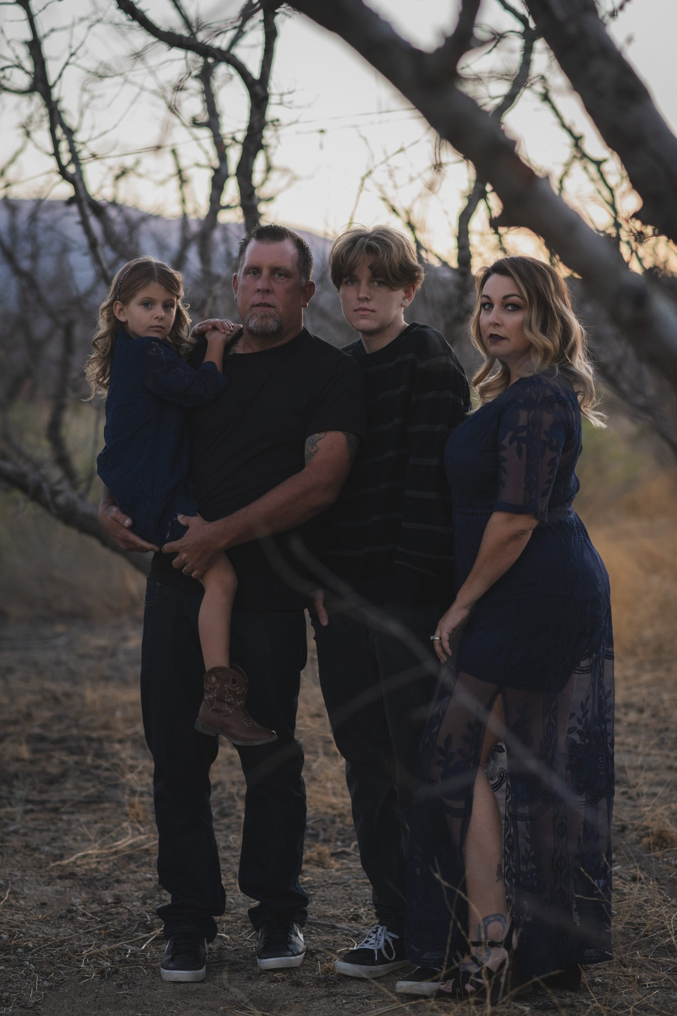 Dark and moody portrait of a family in a dead peach orchard posing for a family photoshoot in the high desert. There are rows of trees and a deep orange sunset setting behind the mountains of the inland empire. The family is wearing dark clothes smiling for the high deserts best family photographer