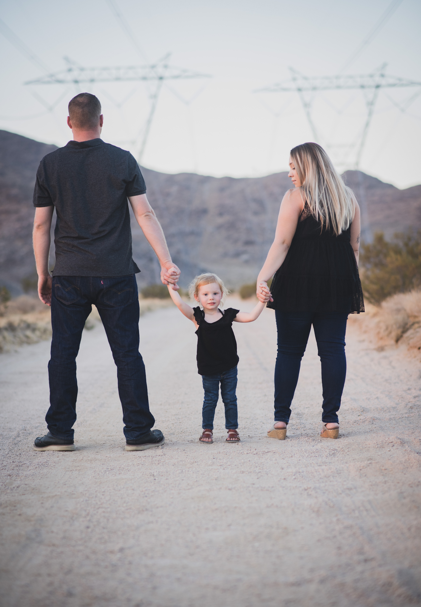 Dark and moody family portraits taken at sunset in the beautiful california mountains in lucerne valley. The high desert sky is behind the mother and father who are wearing red. The three year old daughter is smiling at the high deserts best family photographer