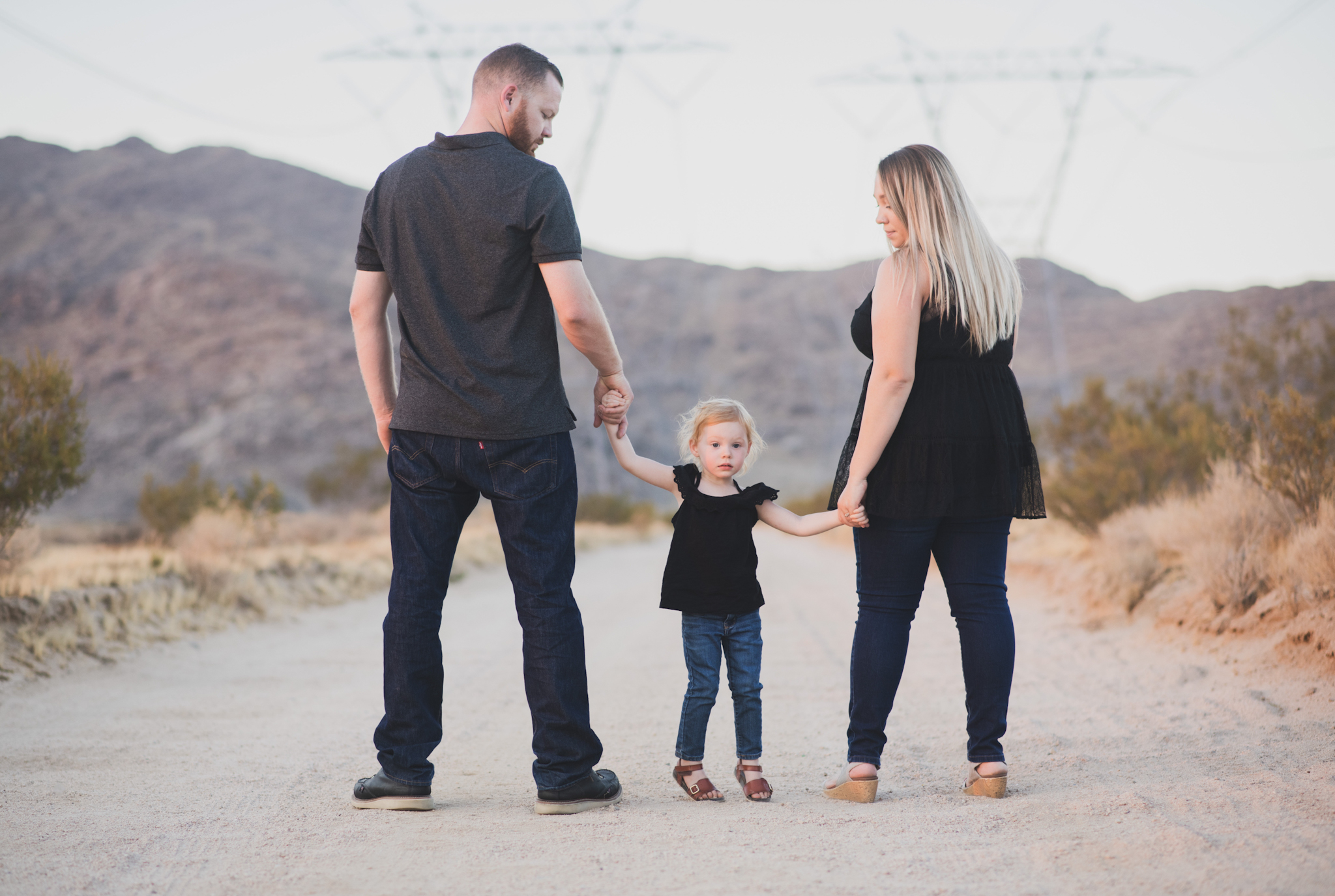 Dark and moody family portraits taken at sunset in the beautiful california mountains in lucerne valley. The high desert sky is behind the mother and father who are wearing red. The three year old daughter is smiling at the high deserts best family photographer