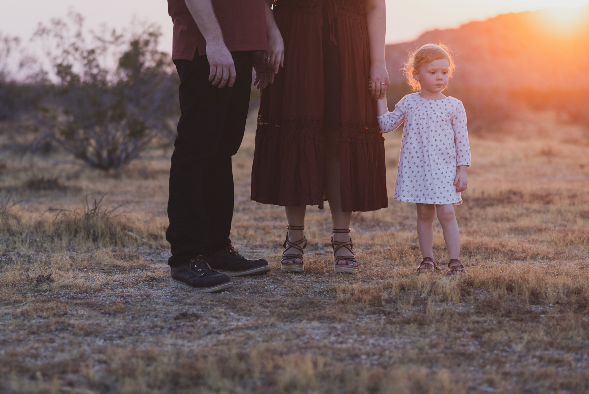 Dark and moody family portraits taken at sunset in the beautiful california mountains in lucerne valley. The high desert sky is behind the mother and father who are wearing red. The three year old daughter is smiling at the high deserts best family photographer
