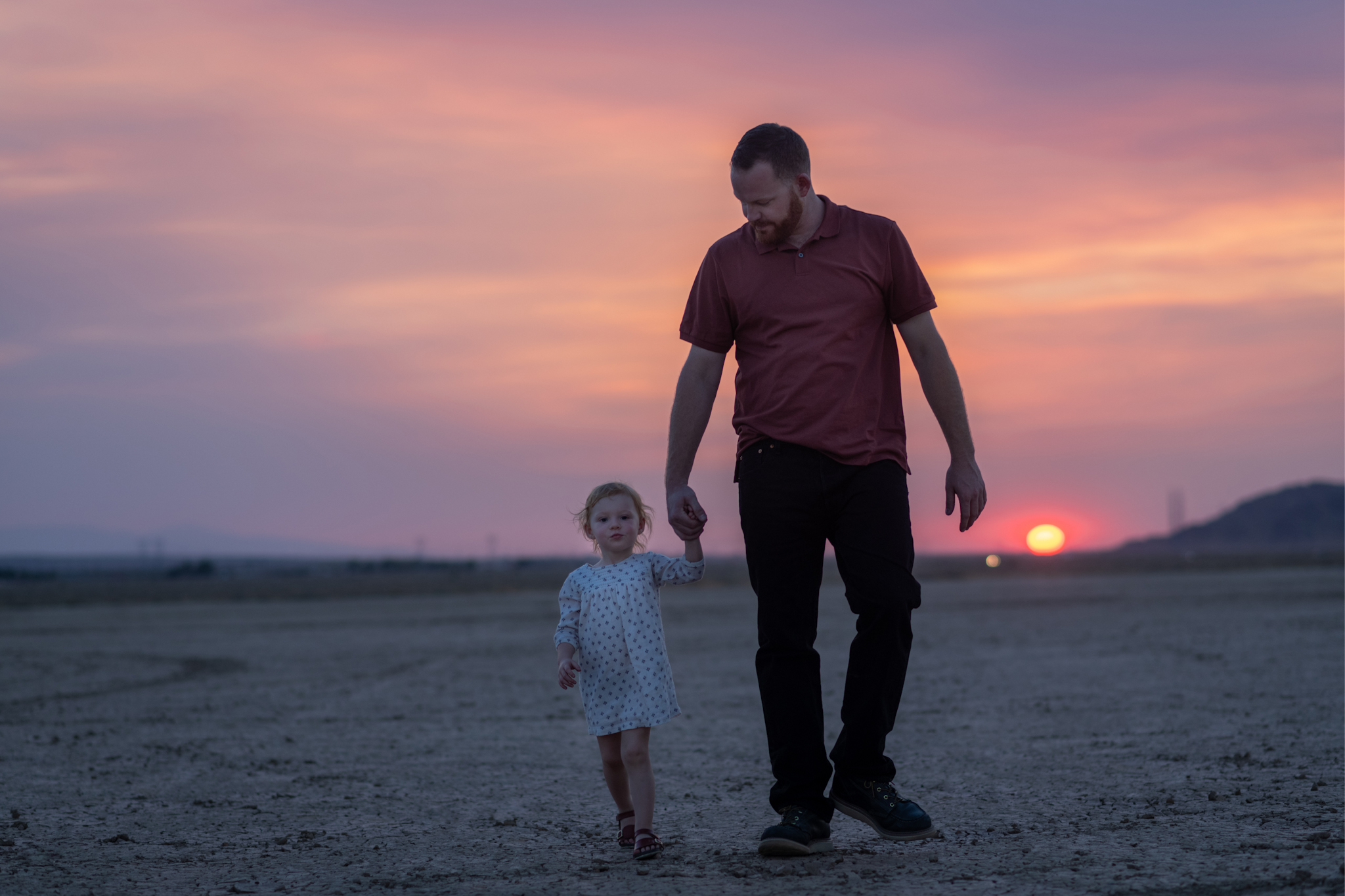 Dark and moody family portraits taken at sunset in the beautiful california mountains in lucerne valley. The high desert sky is behind the mother and father who are wearing red. The three year old daughter is smiling at the high deserts best family photographer