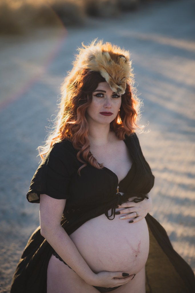 Dark and moody pictures of a pregnant woman with red hair in a black dress posing for the number one photographer in the high desert. She is in a dead peach orchard lined with row of dark bare peach trees. The orange sun is setting in the san bernardino mountains