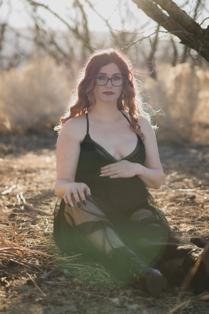 Dark and moody pictures of a pregnant woman with red hair in a black dress posing for the number one photographer in the high desert. She is in a dead peach orchard lined with row of dark bare peach trees. The orange sun is setting in the san bernardino mountains