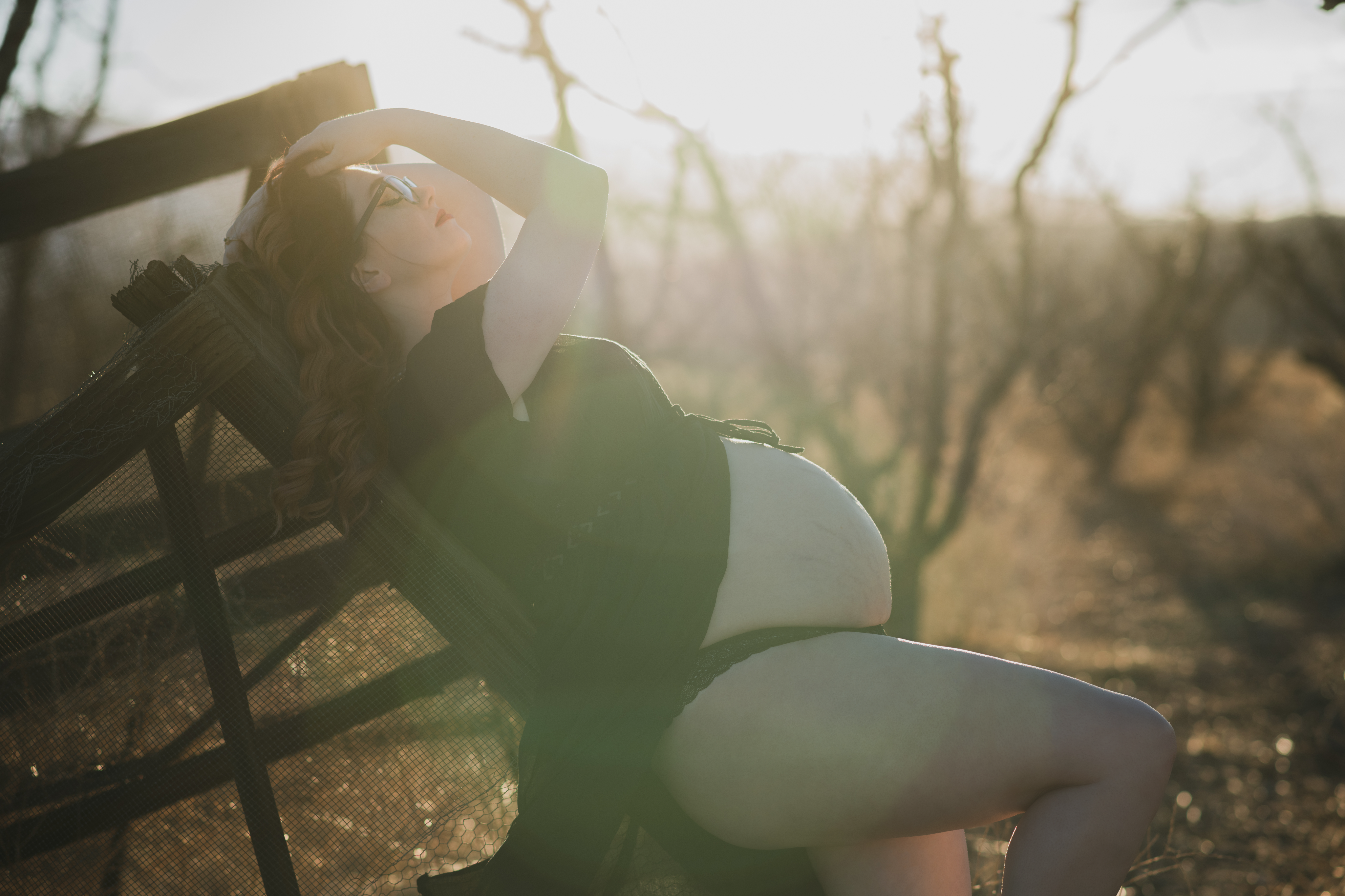 Dark and moody pictures of a pregnant woman with red hair in a black dress posing for the number one photographer in the high desert. She is in a dead peach orchard lined with row of dark bare peach trees. The orange sun is setting in the san bernardino mountains