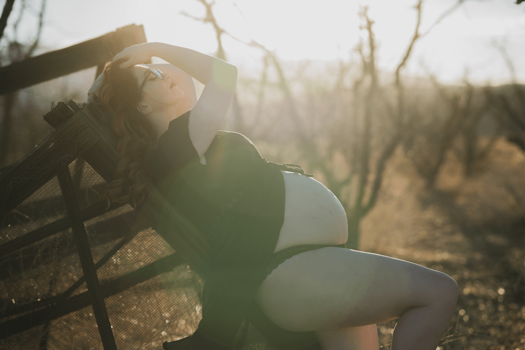 Dark and moody pictures of a pregnant woman with red hair in a black dress posing for the number one photographer in the high desert. She is in a dead peach orchard lined with row of dark bare peach trees. The orange sun is setting in the san bernardino mountains