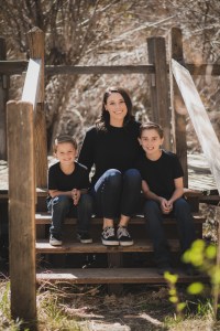 Light and airy portrait of a small family posing for the inland empires best photographer in the green mountains of Wrightwood California. The mother is wearing a white dress with flowers and her two sons are wearing blue shirts and smiling for the camera.