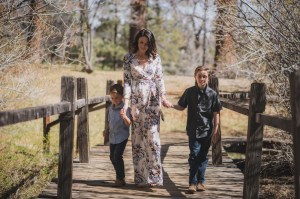 Light and airy portrait of a small family posing for the inland empires best photographer in the green mountains of Wrightwood California. The mother is wearing a white dress with flowers and her two sons are wearing blue shirts and smiling for the camera