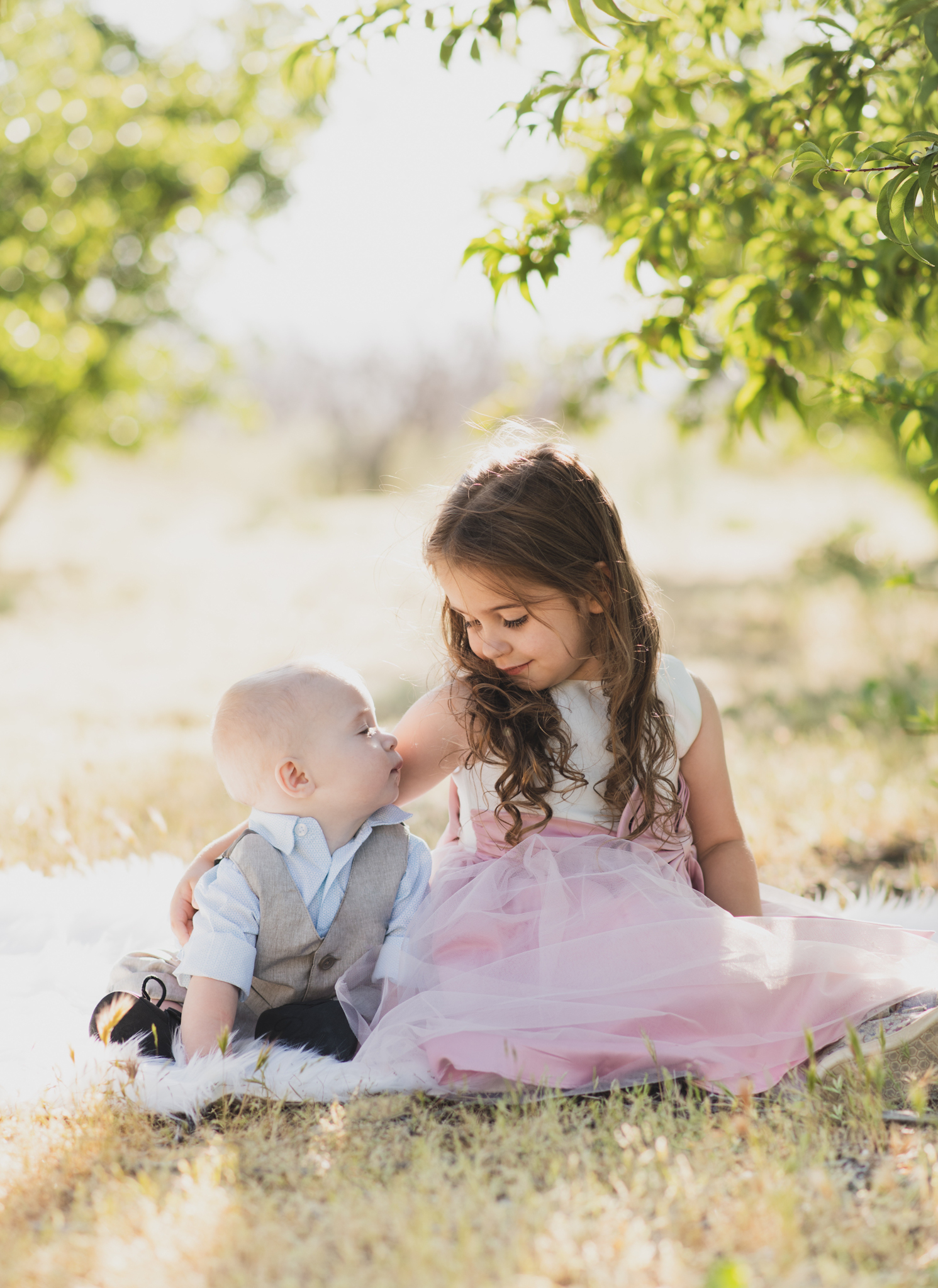 Light and airy portrait of a family in a peach orchard in the high desert. They are smiling for the high deserts best family photographer. the mother is wearing a white dress around green trees. The father and son both are wearing blue shirts and grey vests. The four year old daughter dances and spins for the camera in the inland empire