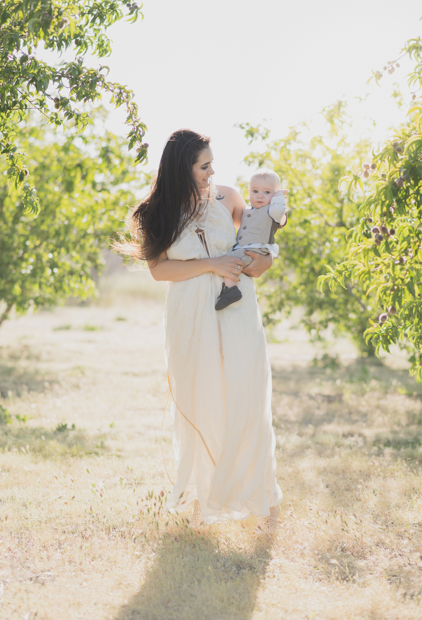 Light and airy portrait of a family in a peach orchard in the high desert. They are smiling for the high deserts best family photographer. the mother is wearing a white dress around green trees. The father and son both are wearing blue shirts and grey vests. The four year old daughter dances and spins for the camera in the inland empire