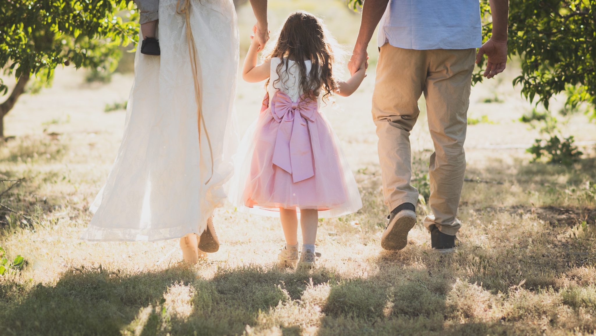 Light and airy portrait of a family in a peach orchard in the high desert. They are smiling for the high deserts best family photographer. the mother is wearing a white dress around green trees. The father and son both are wearing blue shirts and grey vests. The four year old daughter dances and spins for the camera in the inland empire
