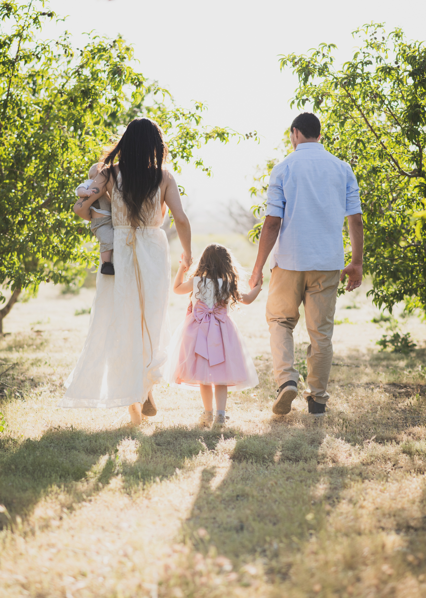 Light and airy portrait of a family in a peach orchard in the high desert. They are smiling for the high deserts best family photographer. the mother is wearing a white dress around green trees. The father and son both are wearing blue shirts and grey vests. The four year old daughter dances and spins for the camera in the inland empire