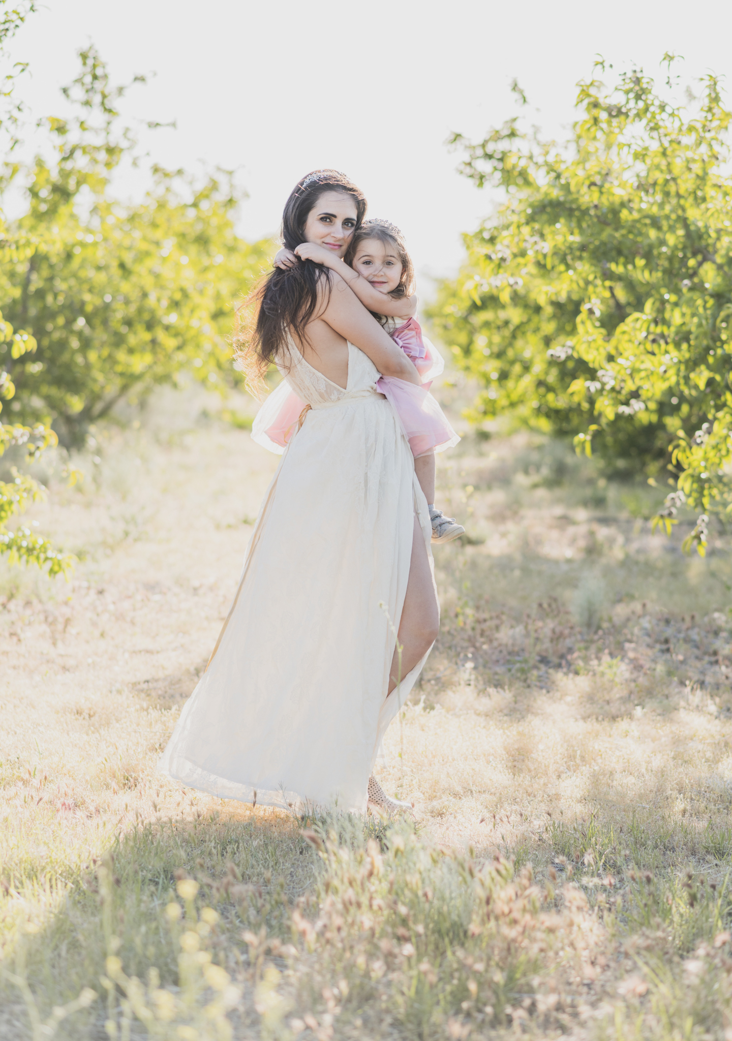 Light and airy portrait of a family in a peach orchard in the high desert. They are smiling for the high deserts best family photographer. the mother is wearing a white dress around green trees. The father and son both are wearing blue shirts and grey vests. The four year old daughter dances and spins for the camera in the inland empire