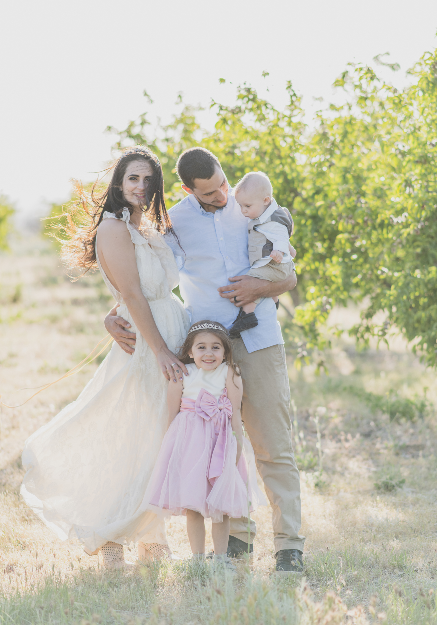 Light and airy portrait of a family in a peach orchard in the high desert. They are smiling for the high deserts best family photographer. the mother is wearing a white dress around green trees. The father and son both are wearing blue shirts and grey vests. The four year old daughter dances and spins for the camera in the inland empire