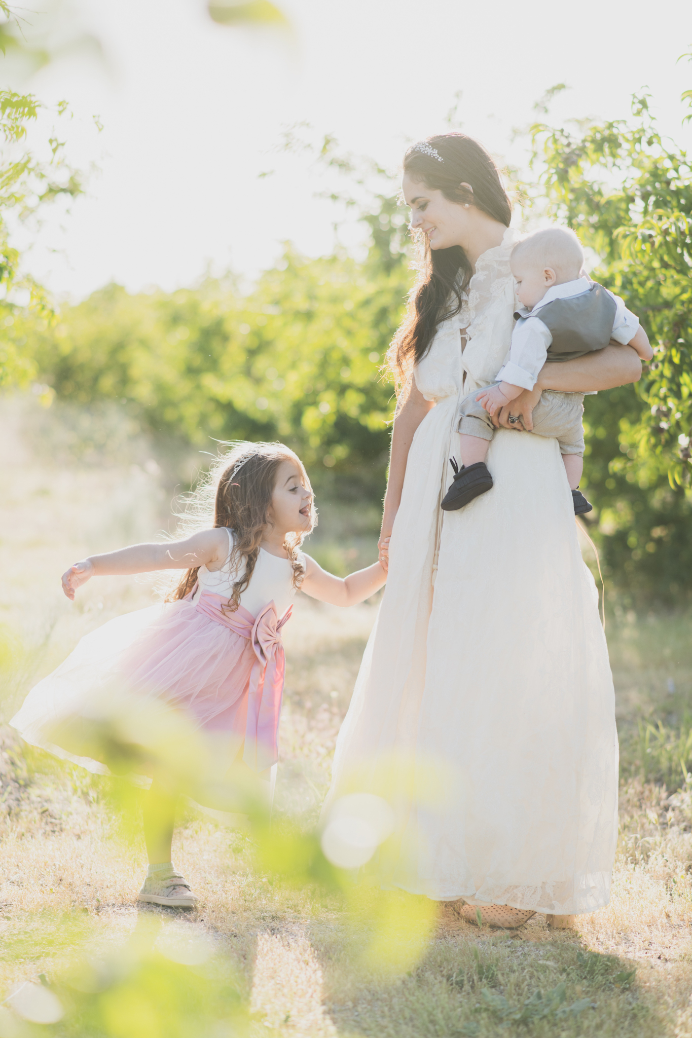 Light and airy portrait of a family in a peach orchard in the high desert. They are smiling for the high deserts best family photographer. the mother is wearing a white dress around green trees. The father and son both are wearing blue shirts and grey vests. The four year old daughter dances and spins for the camera in the inland empire