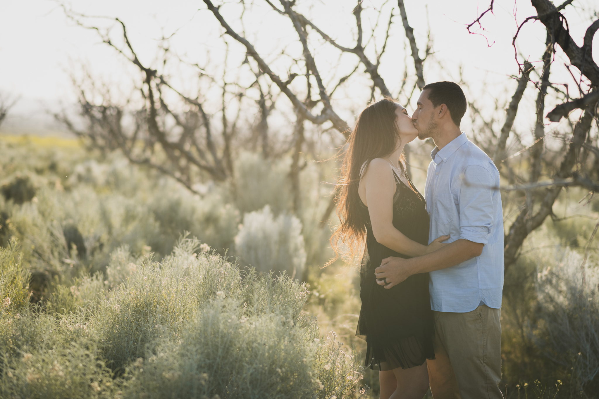 Light and airy portrait of a family in a peach orchard in the high desert. They are smiling for the high deserts best family photographer. the mother is wearing a white dress around green trees. The father and son both are wearing blue shirts and grey vests. The four year old daughter dances and spins for the camera in the inland empire