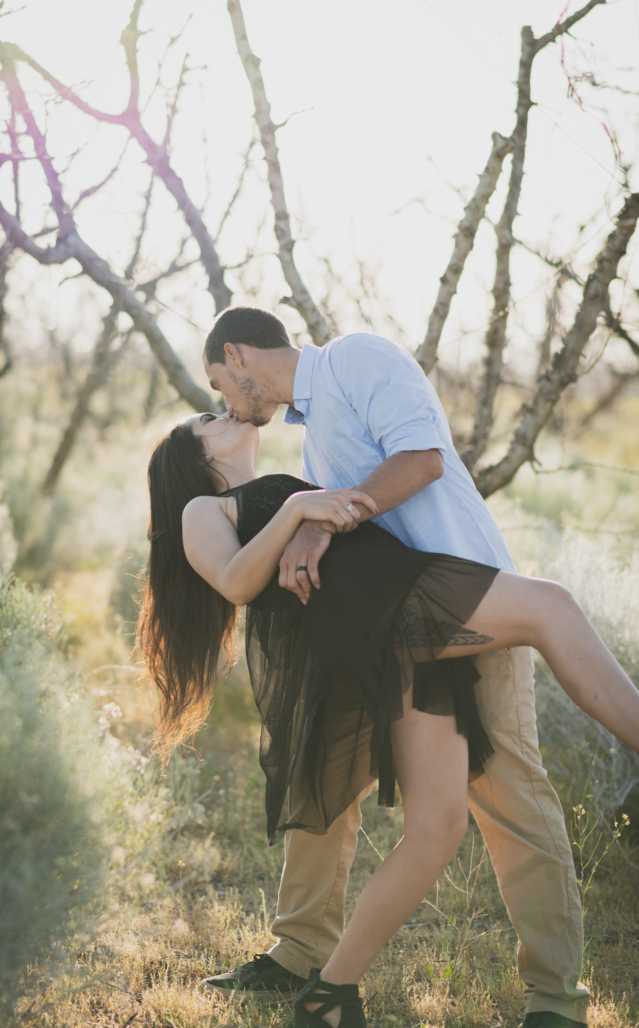 Light and airy portrait of a family in a peach orchard in the high desert. They are smiling for the high deserts best family photographer. the mother is wearing a white dress around green trees. The father and son both are wearing blue shirts and grey vests. The four year old daughter dances and spins for the camera in the inland empire