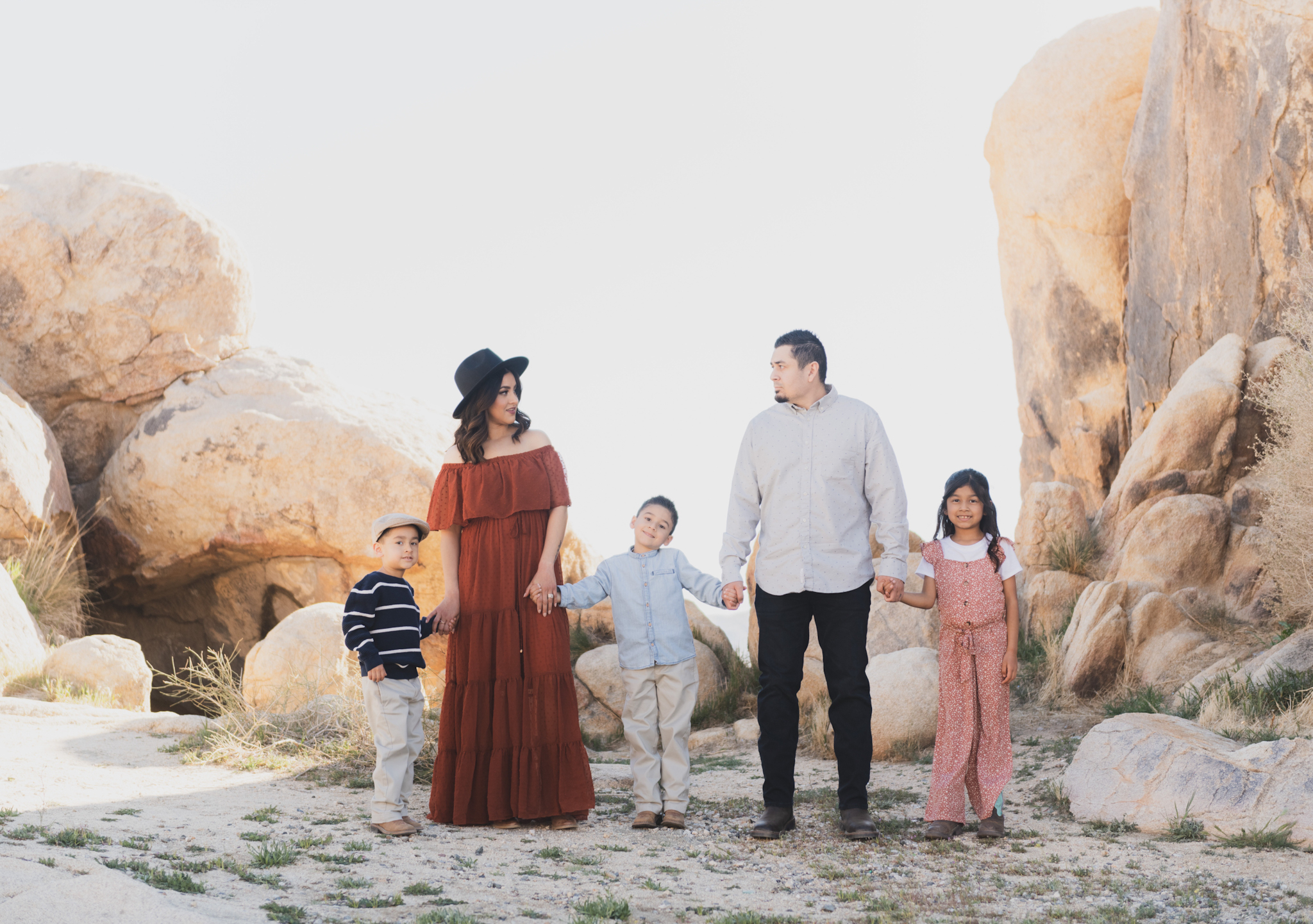 Light and airy portraits of a family in Lucerne Valley California in the High Desert. Three small children are laughing and playing. The desert background is warm with lots of sun, joshua trees and mountains. The mother is wearing a red dress in these inland empire pictures