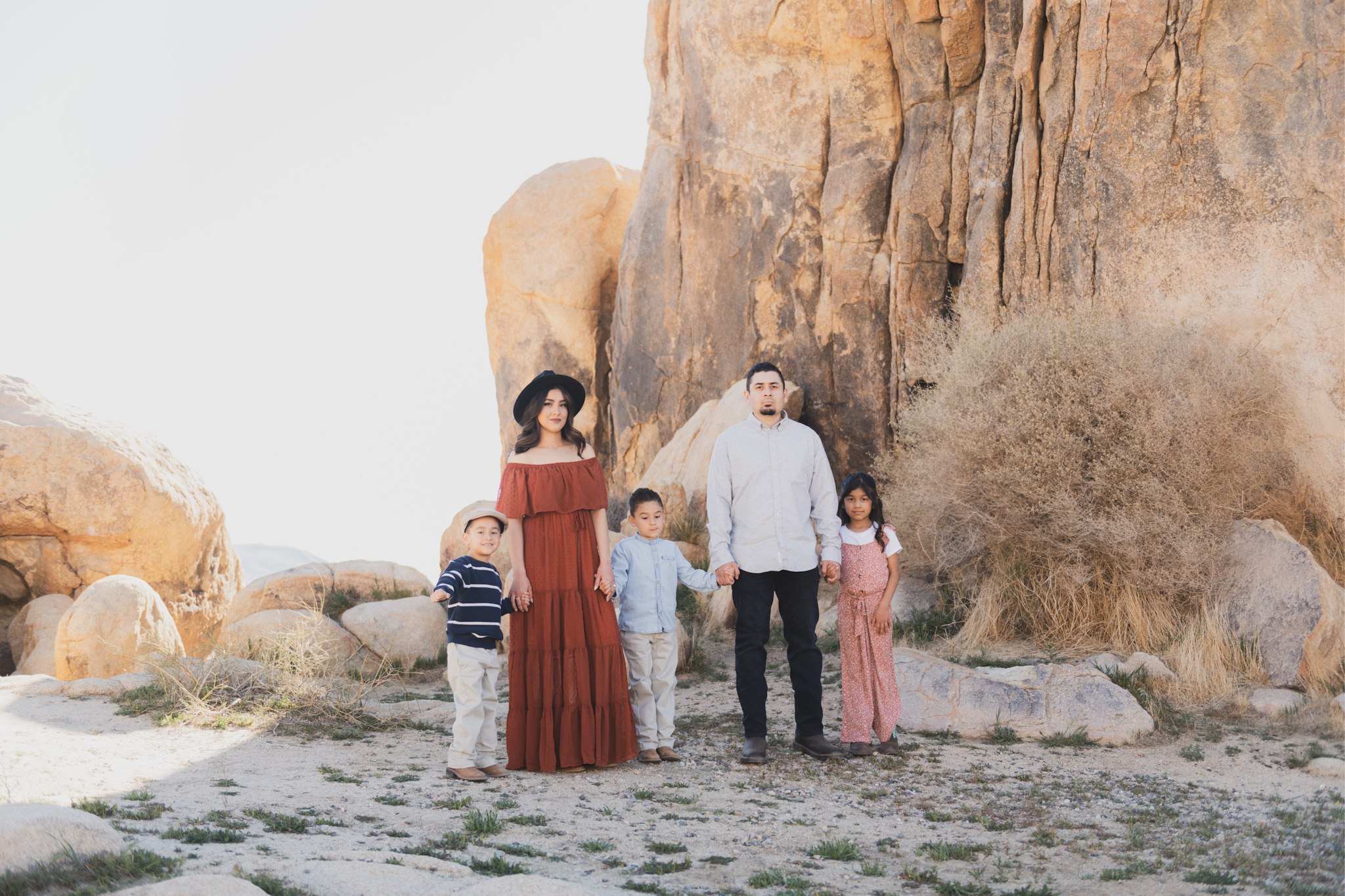 Light and airy portraits of a family in Lucerne Valley California in the High Desert. Three small children are laughing and playing. The desert background is warm with lots of sun, joshua trees and mountains. The mother is wearing a red dress in these inland empire pictures