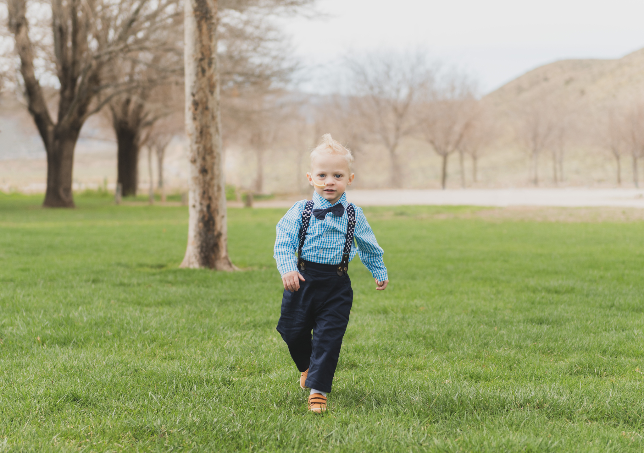 Light and air pictures of a three year old blonde boy playing in the grass at Hesperia Lakes park in Hesperia California in the High desert mountains. Little boy is playing with bubbles and laughing while wearing a blue bow tie and suspenders