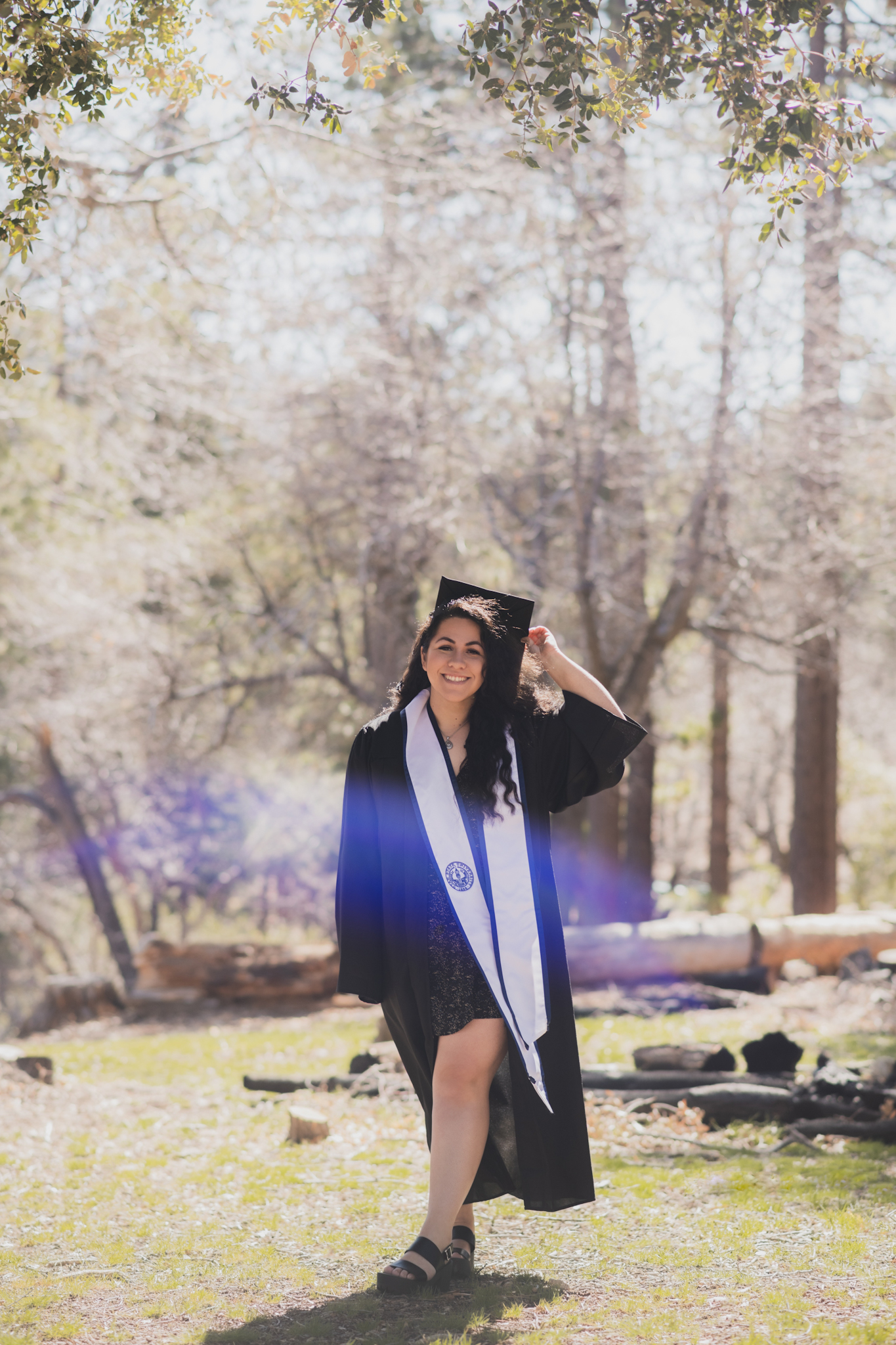 Light and airy portraits of a young woman in a graduation cap and gown. Woman is smiling and happy in the Wrightwood mountains of california. She is happy and throwing her cap in the air surrounded by large trees in these high desert pictures