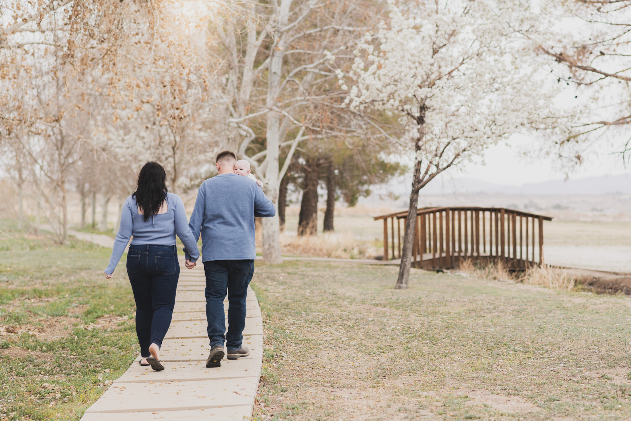 Light and airy pictures of a couple with a baby boy. Couple is wearing blue holding baby boy and smiling in these high desert portraits shot in the beautiful Hesperia Lakes park in the inland empire. There is a tree with white flowers blooming in the background