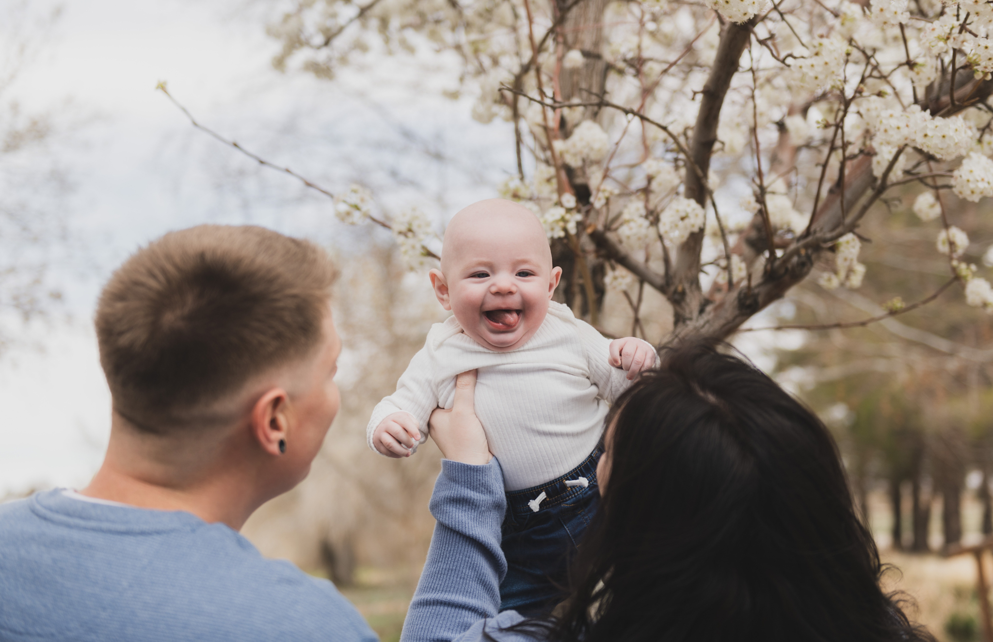 Light and airy pictures of a couple with a baby boy. Couple is wearing blue holding baby boy and smiling in these high desert portraits shot in the beautiful Hesperia Lakes park in the inland empire. There is a tree with white flowers blooming in the background