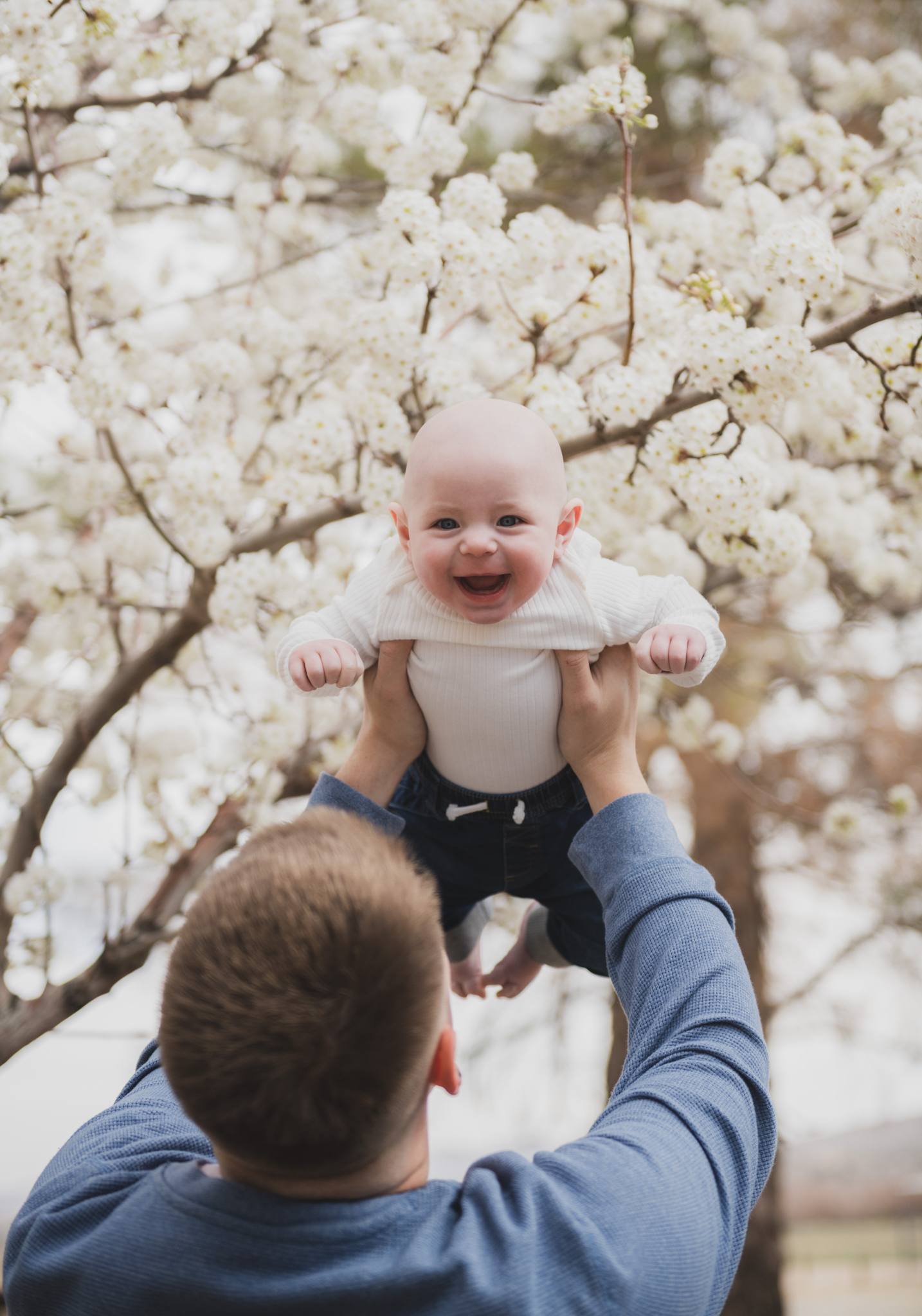 Light and airy pictures of a couple with a baby boy. Couple is wearing blue holding baby boy and smiling in these high desert portraits shot in the beautiful Hesperia Lakes park in the inland empire. There is a tree with white flowers blooming in the background