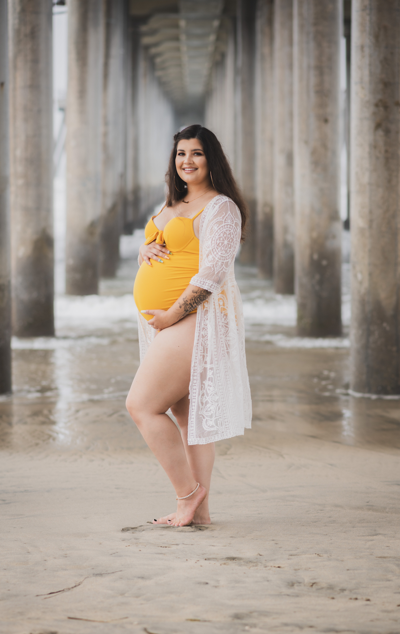 Light and airy portraits of a maternity shoot with family. Pregnant mother is wearing a bright yellow bathing suite in these Huntington Beach pictures. You can see blue waves in the background, the pier and husband and baby boy smiling with mother. Inland empire photoshoot at the beach