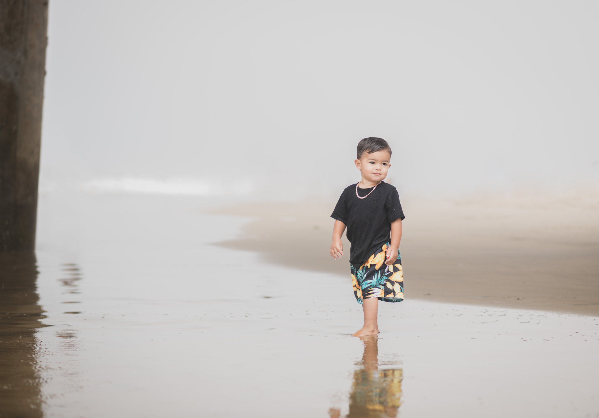 Light and airy portraits of a maternity shoot with family. Pregnant mother is wearing a bright yellow bathing suite in these Huntington Beach pictures. You can see blue waves in the background, the pier and husband and baby boy smiling with mother. Inland empire photoshoot at the beach