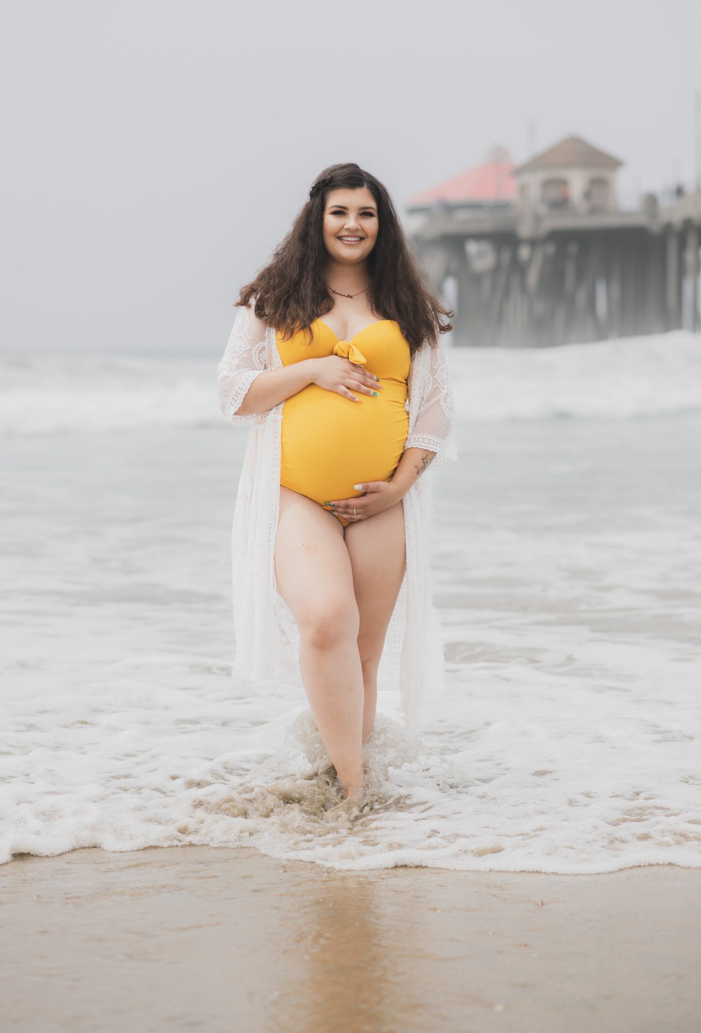Light and airy portraits of a maternity shoot with family. Pregnant mother is wearing a bright yellow bathing suite in these Huntington Beach pictures. You can see blue waves in the background, the pier and husband and baby boy smiling with mother. Inland empire photoshoot at the beach