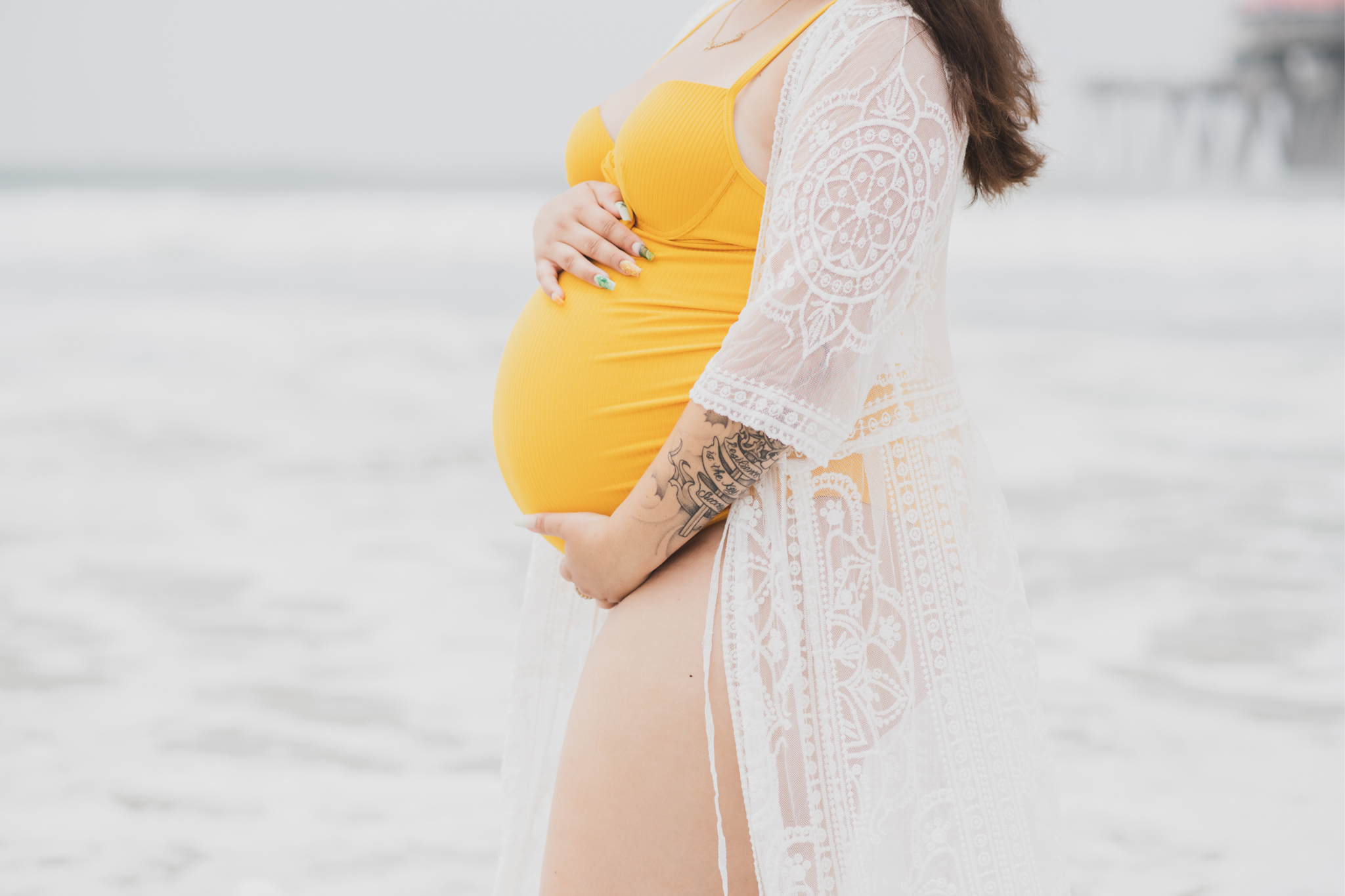 Light and airy portraits of a maternity shoot with family. Pregnant mother is wearing a bright yellow bathing suite in these Huntington Beach pictures. You can see blue waves in the background, the pier and husband and baby boy smiling with mother. Inland empire photoshoot at the beach