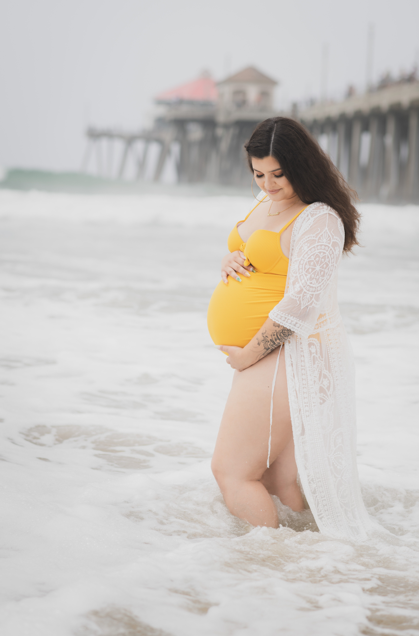 Light and airy portraits of a maternity shoot with family. Pregnant mother is wearing a bright yellow bathing suite in these Huntington Beach pictures. You can see blue waves in the background, the pier and husband and baby boy smiling with mother. Inland empire photoshoot at the beach