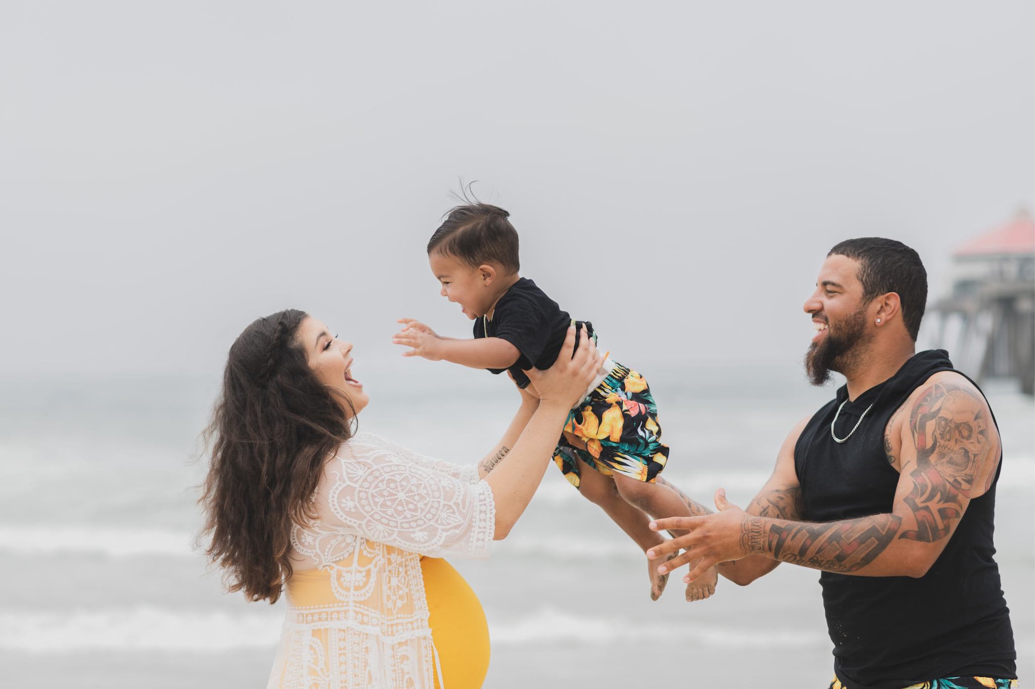 Light and airy portraits of a maternity shoot with family. Pregnant mother is wearing a bright yellow bathing suite in these Huntington Beach pictures. You can see blue waves in the background, the pier and husband and baby boy smiling with mother. Inland empire photoshoot at the beach