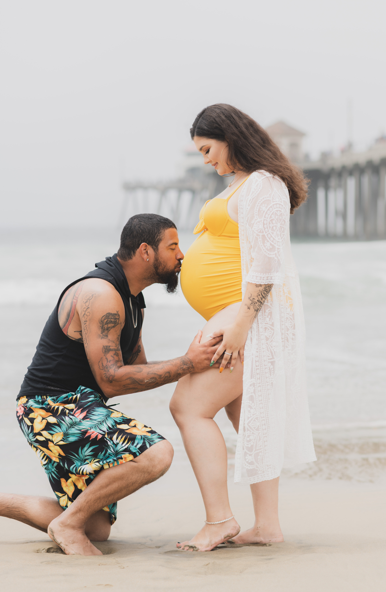 Light and airy portraits of a maternity shoot with family. Pregnant mother is wearing a bright yellow bathing suite in these Huntington Beach pictures. You can see blue waves in the background, the pier and husband and baby boy smiling with mother. Inland empire photoshoot at the beach